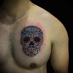 Фото тату на груди: красочный рисунок черепа в стиле Sugar Skull, украшенного сложными узорами и цветами.