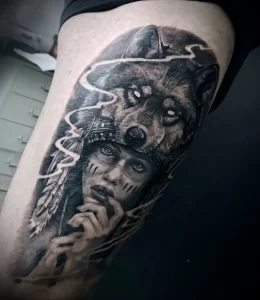 Черно-белая татуировка волка и лица индейца в стиле blackwork на бедре. Детализированный рисунок с использованием геометрических узоров и теней.