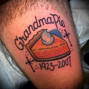Фото тату на предплечье: кусочек тыквенного пирога с надписью "Grandma Pie" и датами "1923-2007", выполненный в стиле олдскул. Памятный рисунок татуировки.