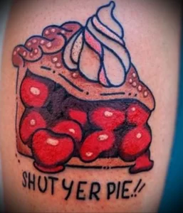 Фото тату: яркий рисунок вишневого пирога с кремом и надписью "SHUT YER PIE!" на руке. Ироничный стиль и оригинальная композиция.