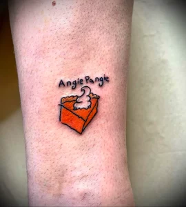 Небольшое фото тату на руке с изображением кусочка пирога и надписью 'Angie Pangie'. Стиль татуировки - минимализм, преобладают оранжевые и черные цвета.
