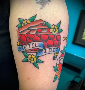 Фото тату на предплечье: классический рисунок пирога с надписью 'Pie Till I Die', украшенный цветами и лентой в стиле олд скул.