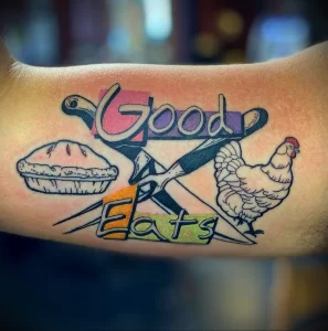 Фото татуировки на руке с изображением пирога, курицы и надписи 'Good Eats'. Яркие цвета и ретро-стиль.