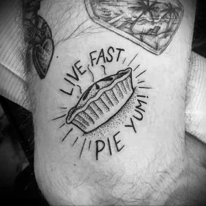 Фото тату на руке с изображением куска пирога в круге с надписью "Live Fast, Pie Yumi". Простой и стильный рисунок татуировки.