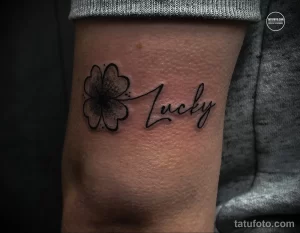 Фото тату четырехлистного клевера и надписи 'Lucky' на предплечье. Черно-белый рисунок татуировки, выполненный в каллиграфическом стиле.