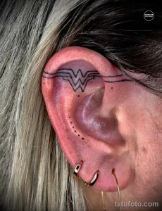 Фото тату на ухе: стилизованное изображение символа Wonder Woman, выполненное в черных линиях и точках. Идеальный выбор для поклонников комиксов.