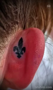 Фото тату: небольшая татуировка с изображением Fleur-de-lis на ухе, выполненная в черном цвете. Лаконичный и элегантный дизайн.