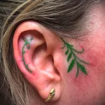 Фото тату: татуировка с изображением Fleur-de-lis (флер-де-лис) на ухе, выполненная в красном цвете. Классический символ с четкими линиями.