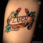 Фото татуировки с надписью 'LUCK' на ленте, окруженной двумя руками, держащими цветы, в стиле олд скул на руке.