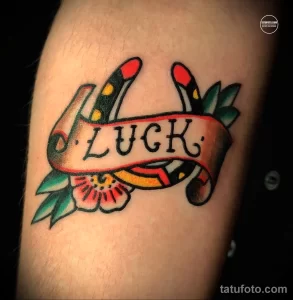 Фото татуировки с надписью 'LUCK' на ленте, окруженной двумя руками, держащими цветы, в стиле олд скул на руке.