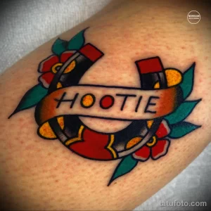 Фото тату на коже: подкова, красная роза и надпись 'HOOTIE' в ярком стиле олд скул. Классические мотивы и насыщенные цвета.