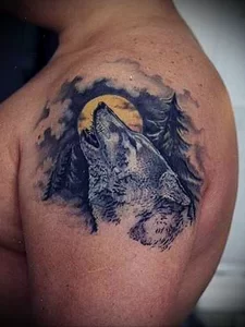 Черно-серая татуировка волка, воющего на луну, на плече. Реалистичный рисунок с детальной проработкой шерсти и фона.