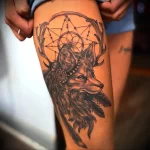 Фото тату на бедре: волк с рогами и ловцом снов, выполненным в технике blackwork. Детализированный рисунок татуировки с перьями и геометрическими элементами.