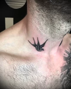 Фото тату маленькой черной ласточки на шее мужчины с щетиной. Классический рисунок татуировки в минималистичном стиле.