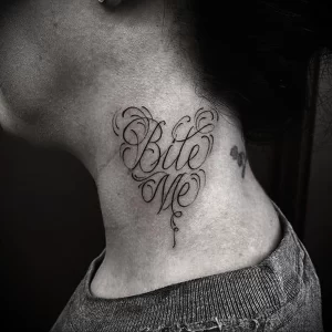 Фото тату: черная татуировка с надписью "Bite Me" в форме сердца на женской шее. Каллиграфический стиль, оригинальный дизайн.