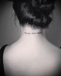 Фото татуировки надписи 'courage, dear heart' на затылке девушки. Черный цвет, минималистичный шрифт, вдохновляющая фраза, идеи для тату.