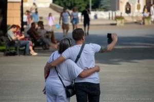 Фотография мужчины с татуировкой на плече, обнимающего девушку на городской улице. Татуировка выполнена в темных тонах, видны детали рисунка. Снимок сделан в солнечный день.