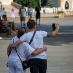 Фотография мужчины с татуировкой на плече, обнимающего девушку на городской улице. Татуировка выполнена в темных тонах, видны детали рисунка. Снимок сделан в солнечный день.