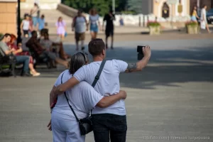 Фотография мужчины с татуировкой на плече, обнимающего девушку на городской улице. Татуировка выполнена в темных тонах, видны детали рисунка. Снимок сделан в солнечный день.