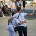 Фотография мужчины с татуировкой на плече, обнимающего девушку на городской улице. Татуировка выполнена в темных тонах, видны детали рисунка. Снимок сделан в солнечный день.