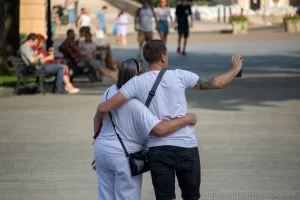 Фотография мужчины с татуировкой на плече, обнимающего девушку на городской улице. Татуировка выполнена в темных тонах, видны детали рисунка. Снимок сделан в солнечный день.
