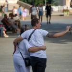 Фотография мужчины с татуировкой на плече, обнимающего девушку на городской улице. Татуировка выполнена в темных тонах, видны детали рисунка. Снимок сделан в солнечный день.