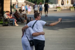 Фотография мужчины с татуировкой на плече, обнимающего девушку на городской улице. Татуировка выполнена в темных тонах, видны детали рисунка. Снимок сделан в солнечный день.