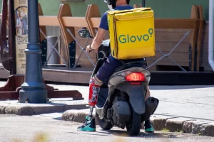 Фото курьера Glovo на скутере с татуировкой на ноге, снятое сбоку. Курьер одет в синюю форму и белый шлем, на спине большой желтый бокс Glovo.