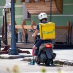Фото курьера Glovo на скутере с татуировкой на ноге, снятое сзади. Курьер одет в синюю форму и белый шлем, на спине большой желтый бокс Glovo.