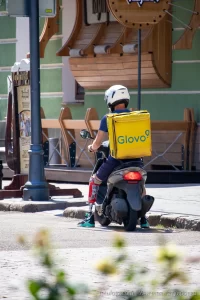 Фото курьера Glovo на скутере с татуировкой на ноге, снятое сзади. Курьер одет в синюю форму и белый шлем, на спине большой желтый бокс Glovo.