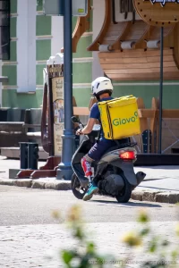 Фото курьера Glovo на скутере с татуировкой на ноге. Курьер одет в синюю форму и белый шлем.