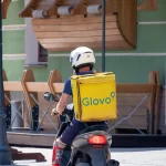Фото курьера Glovo на скутере с татуировкой на ноге, снятое сзади. Курьер одет в синюю форму и белый шлем, на спине большой желтый бокс Glovo.