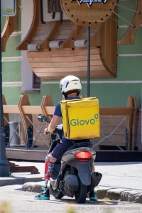 Фото курьера Glovo на скутере с татуировкой на ноге, снятое сзади. Курьер одет в синюю форму и белый шлем, на спине большой желтый бокс Glovo.