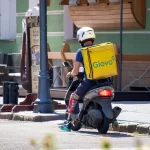 Фото курьера Glovo на скутере с татуировкой на ноге. Курьер одет в синюю форму и белый шлем.