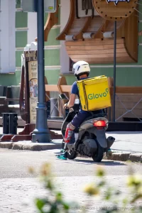 Фото курьера Glovo на скутере с татуировкой на ноге. Курьер одет в синюю форму и белый шлем.