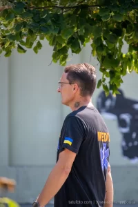 Фото тату: небольшая черно-серая татуировка в виде абстрактного узора на шее мужчины. Минималистичный рисунок с четкими линиями.