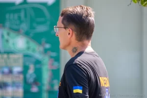 Фото тату: небольшая черно-серая татуировка в виде абстрактного узора на шее мужчины. Минималистичный рисунок с четкими линиями.
