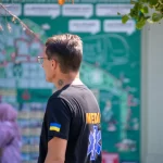 Фото тату: небольшая черно-серая татуировка в виде абстрактного узора на шее мужчины. Минималистичный рисунок с четкими линиями.