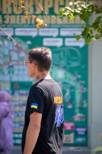 Фото тату: небольшая черно-серая татуировка в виде абстрактного узора на шее мужчины. Минималистичный рисунок с четкими линиями.