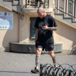 Фото мужчины с татуировкой на руке и футболкой с изображением черепа, идущего по улице.