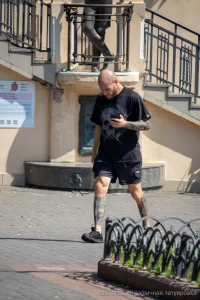 Фото мужчины с татуировкой на руке и футболкой с изображением черепа, идущего по улице.