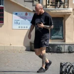 Фото мужчины с татуировками на руках и ногах, идущего по улице и разговаривающего по телефону. Видны детализированные рисунки в темных тонах.