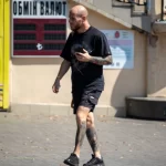 Фото мужчины с татуировками на руках и ногах, идущего по улице и разговаривающего по телефону. Видны детализированные рисунки в темных тонах.