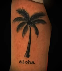 Фото татуировки с надписью 'Aloha' и изображением пальмы на руке. Черно-белый рисунок, тропический стиль, популярные идеи для тату.