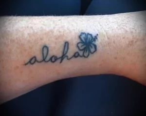 Фото тату надписи 'Aloha' с изображением цветка гибискуса. Изящные линии и нежные цвета создают романтичное и тропическое настроение.