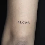 Фото тату надписи 'Aloha' выполненной тонким шрифтом на коже. Простой и элегантный дизайн, подчеркивающий любовь к гавайской культуре.