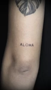 Фото тату надписи 'Aloha' выполненной тонким шрифтом на коже. Простой и элегантный дизайн, подчеркивающий любовь к гавайской культуре.