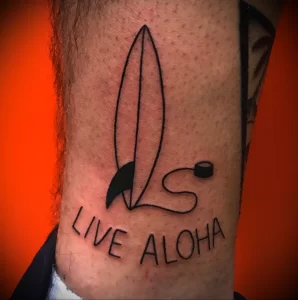 Фото тату с изображением серфа и надписи 'Live Aloha'. Простой, но стильный дизайн, выполненный в черном цвете, подчеркивает любовь к серфингу и гавайской культуре.