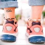 Фото татуировки надписи 'Aloha' на лодыжке. Черно-белое изображение на коже, вид снизу с джинсами.