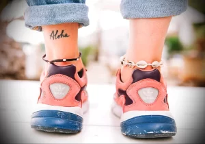 Фото татуировки надписи 'Aloha' на лодыжке. Черно-белое изображение на коже, вид снизу с джинсами.